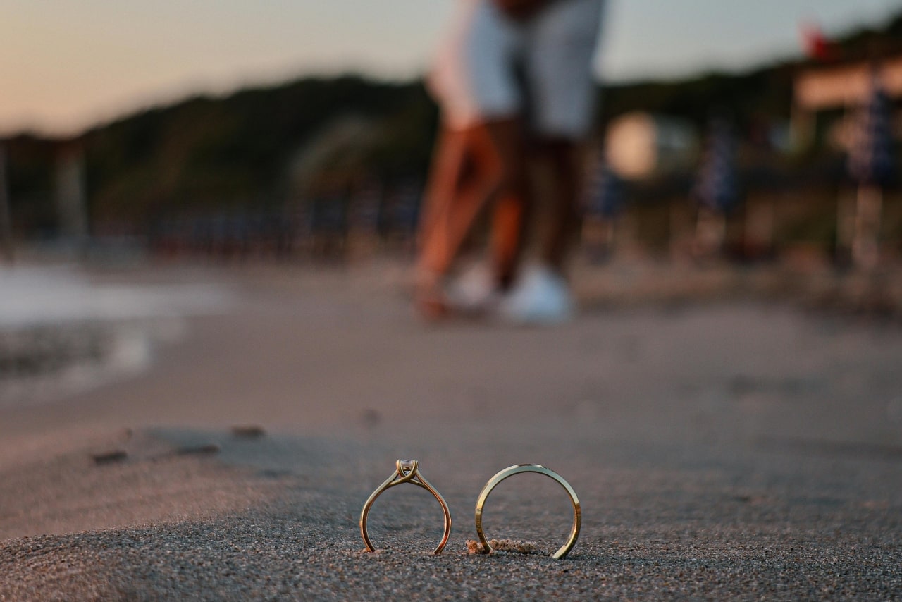 a close up of an engagement ring and wedding band set that is standing up in the sand at the beach with a couple out of focus in the background kissing after the sunset proposal