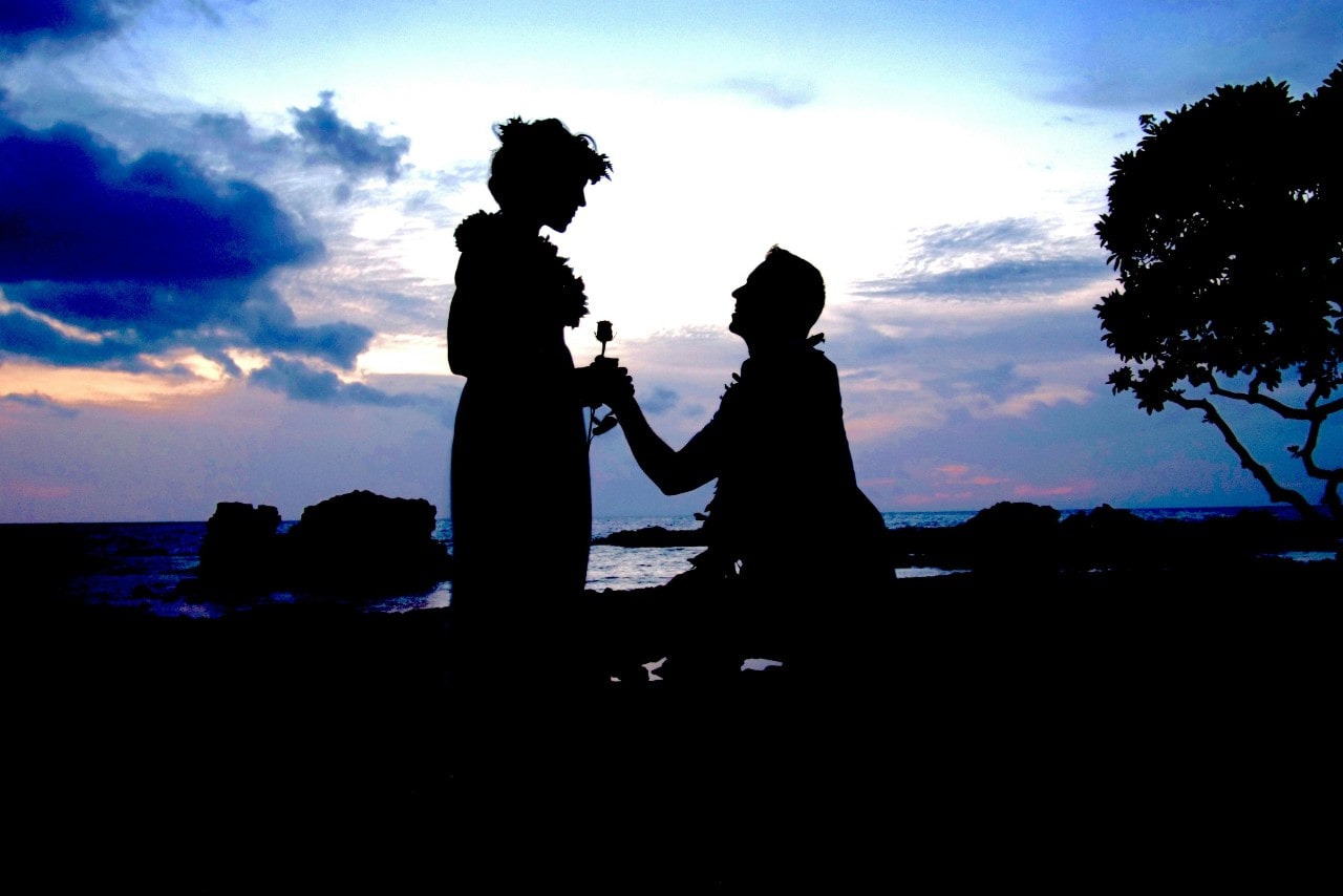 A backlit photo of a man on one knee proposing to his partner at sunrise.