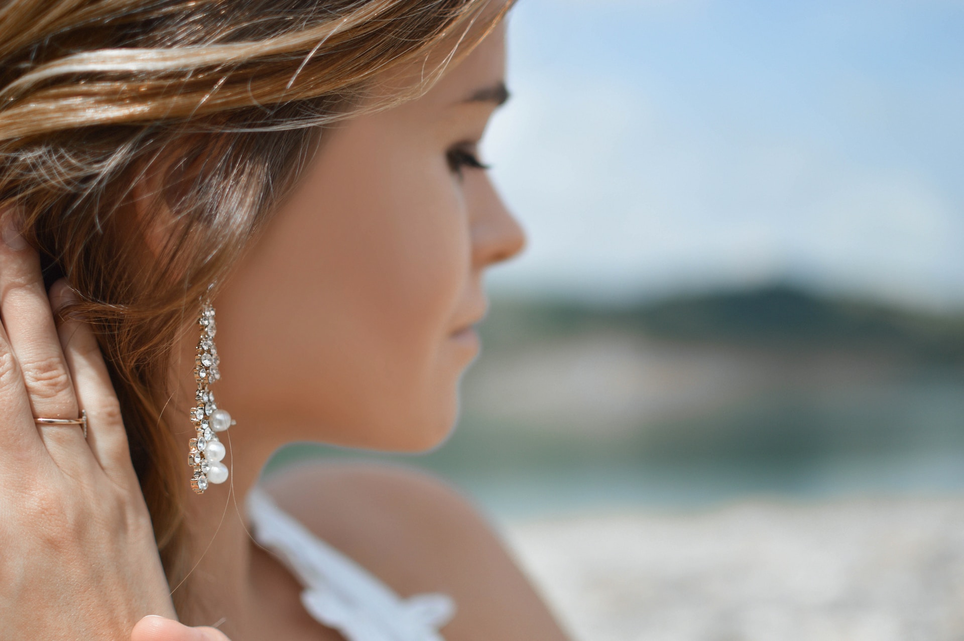 a woman looking away from the camera wearing white gold earrings with a beach in the background