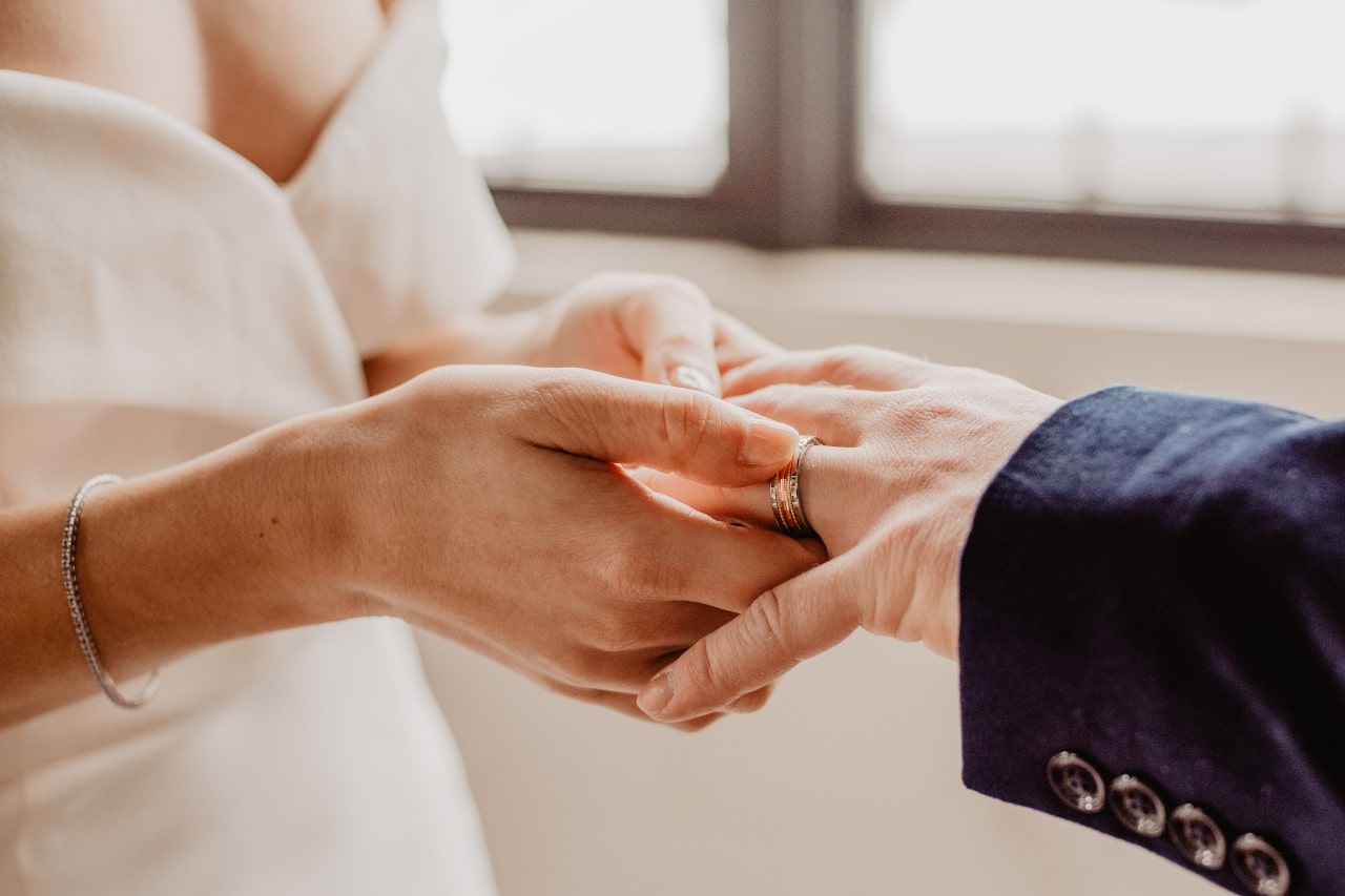 A bride placing a wide wedding band on her groom’s finger.